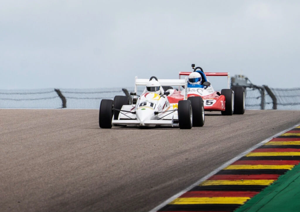Ein weißer Formel-Rennwagen (Startnummer 61) führt vor einem rot-weißen Formelwagen über eine Kuppe der Rennstrecke.