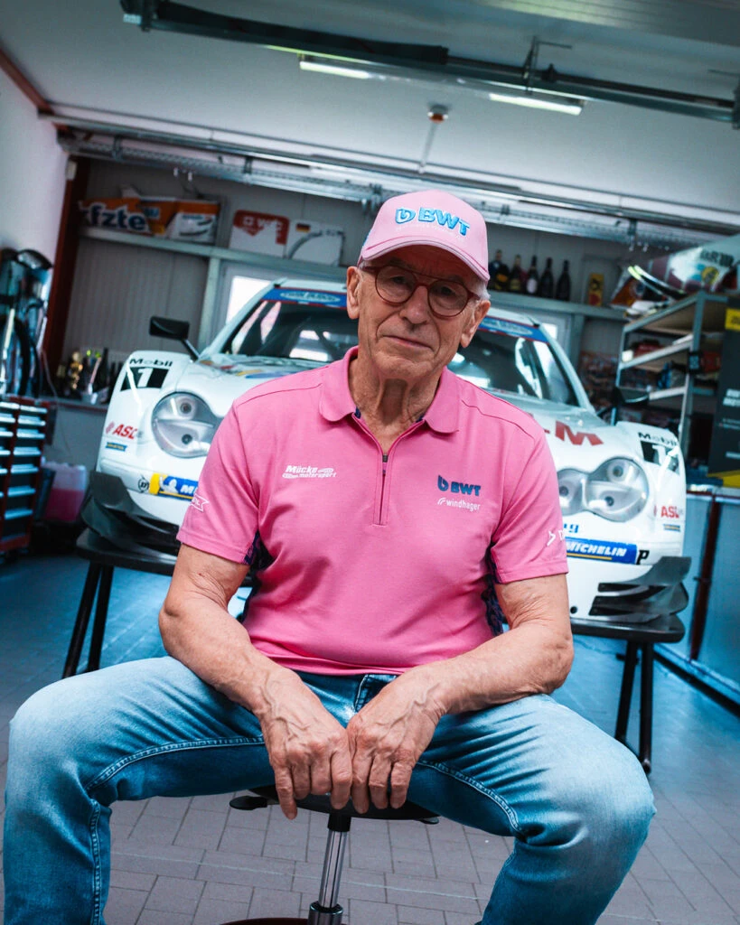 Motorsport-Ikone Peter Mücke von Mücke Motorsport sitzt in Pink gekleidet vor einem Rennwagen in seiner Werkstatt in Berlin