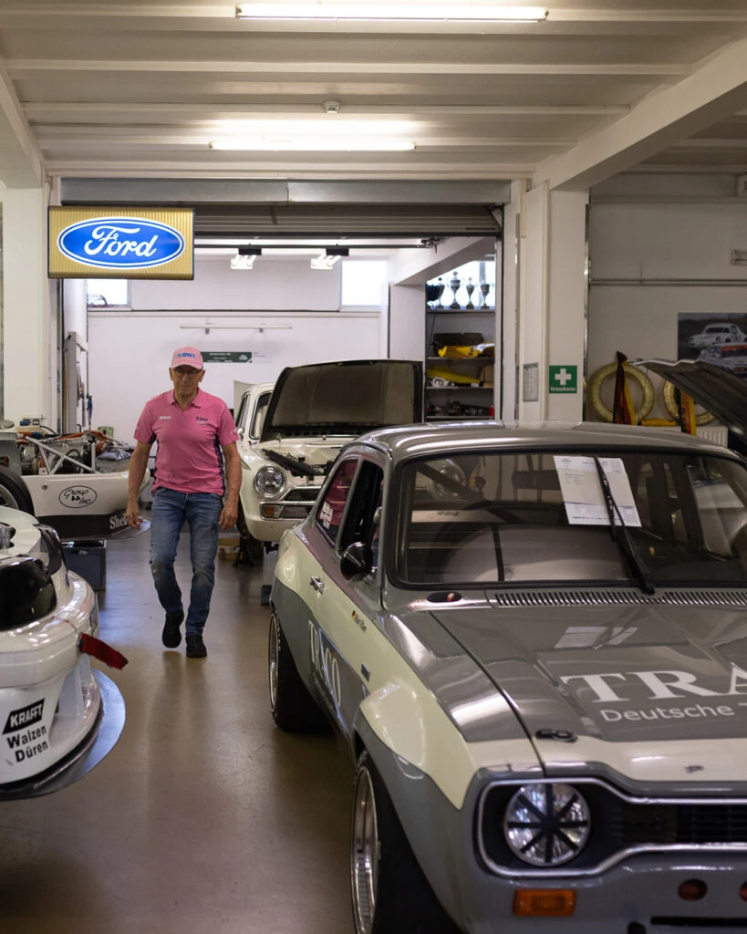 Peter Mücke geht durch seine Werkstatt in Berlin, vorbei an klassischen Rennwagen wie einem Ford Escort.