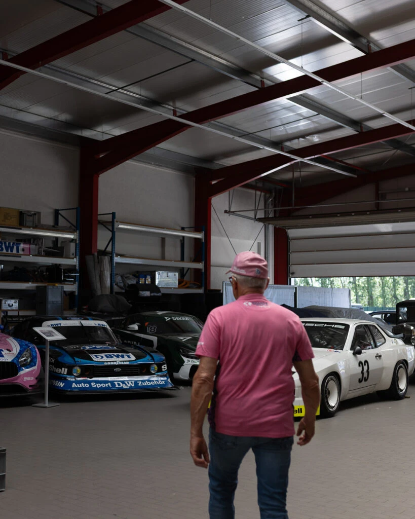 Peter Mücke von hinten in einer großen Halle von Mücke Motorsport, umgeben von verschiedenen Rennwagen.