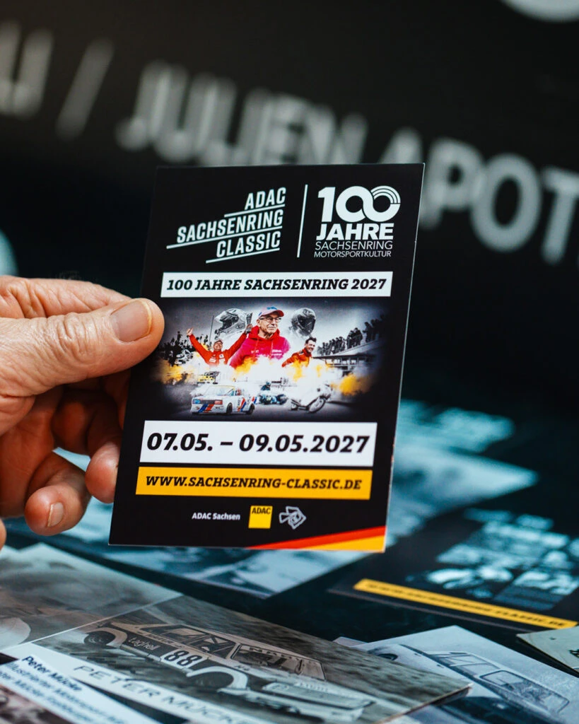 Nahaufnahme einer Hand, die einen Flyer für das Event 100 Jahre Sachsenring Classic 2027 hält.