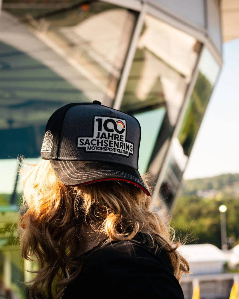 Frau mit blonden Haaren trägt die 100 Jahre Sachsenring Motorsportkultur Cap an der Rennstrecke.